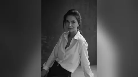 Elegant woman in a white button-down shirt posing in a moody black and white studio portrait.