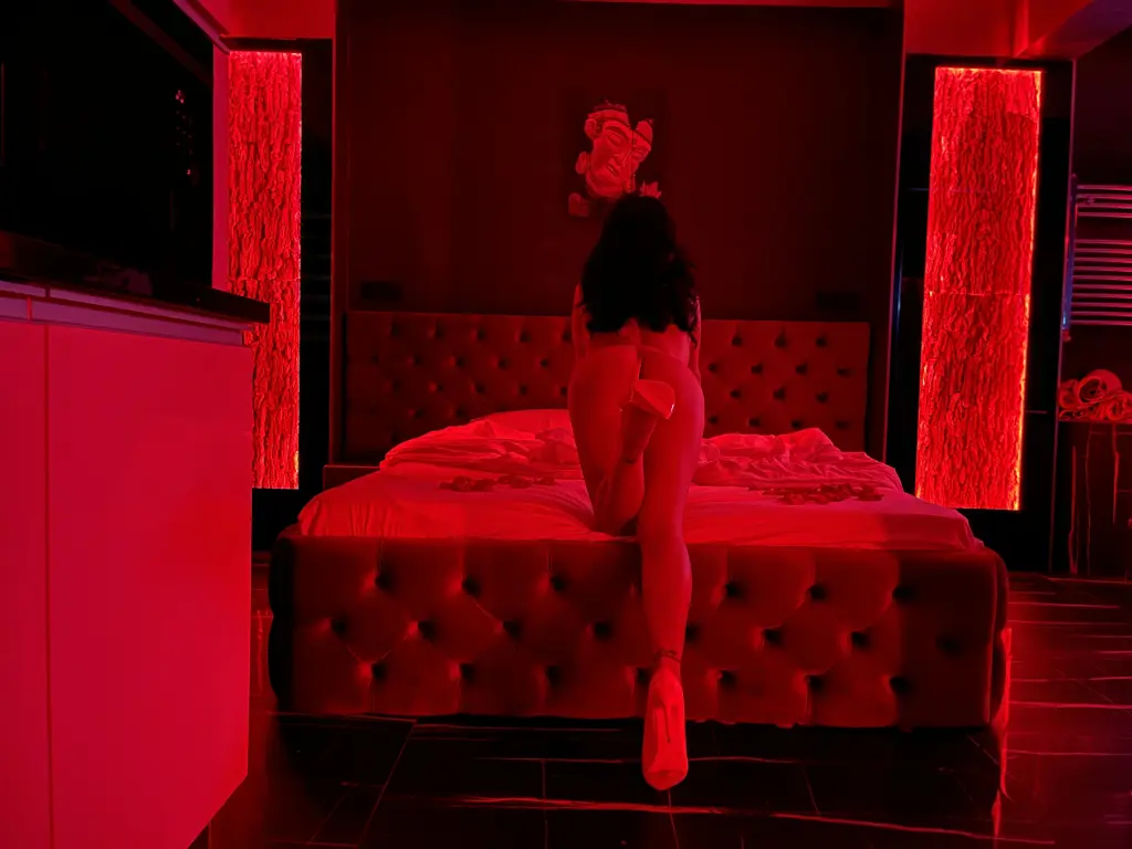 KimRiver — Nude woman sitting on a bed in a moody, red-lit bedroom setting.