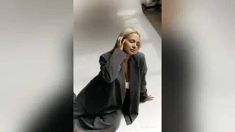 Blonde woman in a grey oversized blazer posing thoughtfully in a minimalist white studio setting.