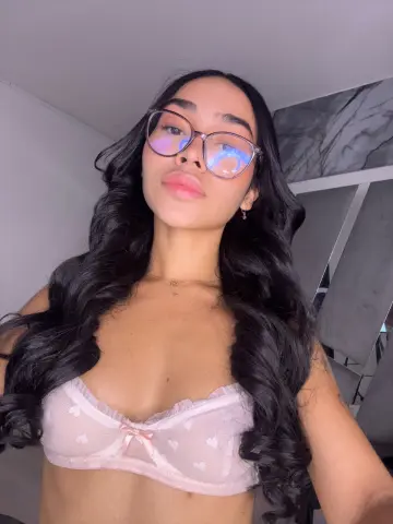 Young woman with long wavy hair wearing glasses and a pink lace bralette in a modern indoor setting.