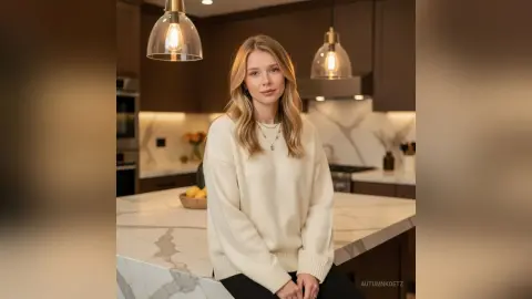 Blonde woman in a cozy cream sweater sitting in a modern luxury kitchen with warm lighting.