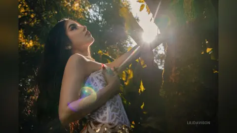 Dreamy portrait of a woman in a white patterned top posing in sunlit forest greenery.