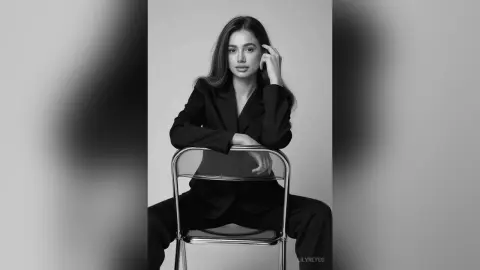 Elegant woman in a black blazer sitting on a chair in a minimalist black and white studio portrait.