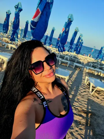 Smiling woman in a purple tank top relaxing on a sunny beach under blue umbrellas.