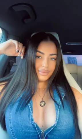 Young woman with long dark hair wearing a blue denim top sitting in a car on a sunny day.