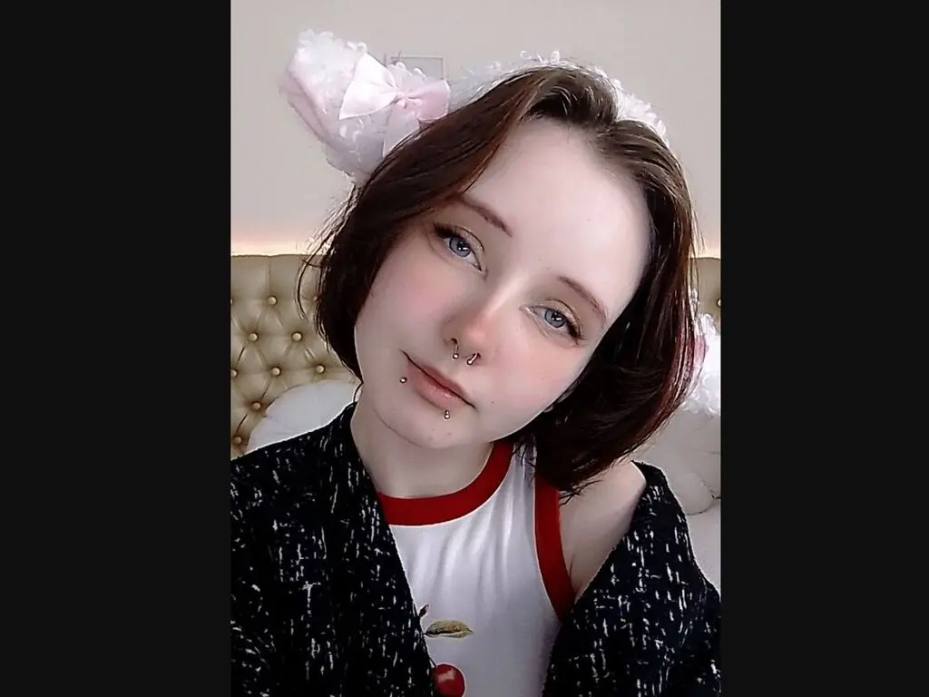 Young woman with short brown hair and nose piercing wearing a white t-shirt and black cardigan in a cozy bedroom.