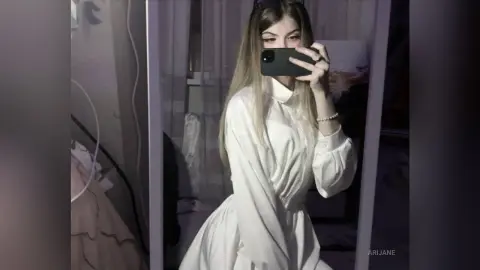 Young woman with long blonde hair wearing a white long-sleeve satin dress taking a mirror selfie in a bedroom.