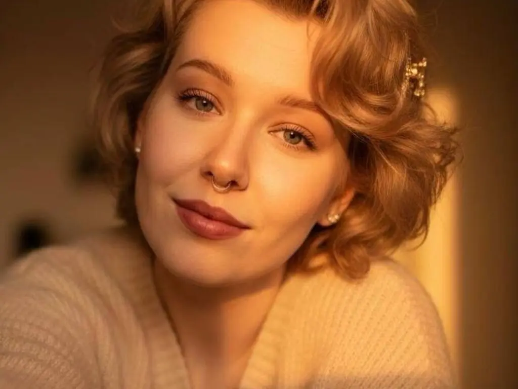 IngritEgos poses close up with wavy blonde hair and a nose ring, wearing a white knit sweater in warm lighting.