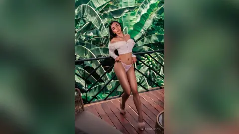 Glamorous woman in a white off-the-shoulder crop top and bikini posing on a tropical balcony surrounded by lush greenery.