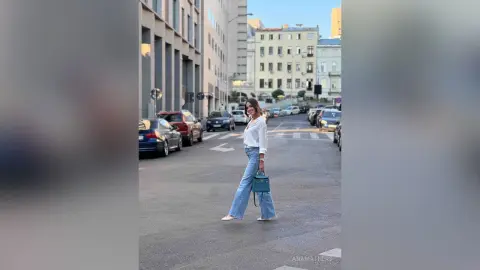 AnaMathers stands on a city street wearing a white long-sleeve shirt and light blue flared jeans, holding a small blue handbag.