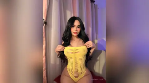 Young woman with long dark hair wearing a yellow cutout bodysuit in a bright indoor setting.