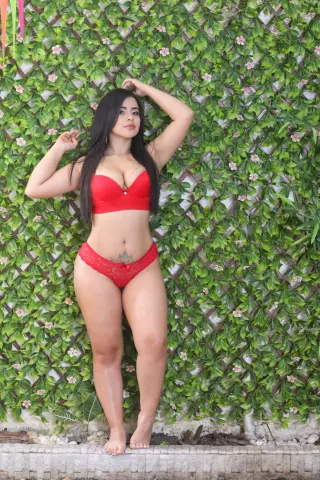Curvy woman in red lace lingerie posing confidently against a lush green leaf wall background.