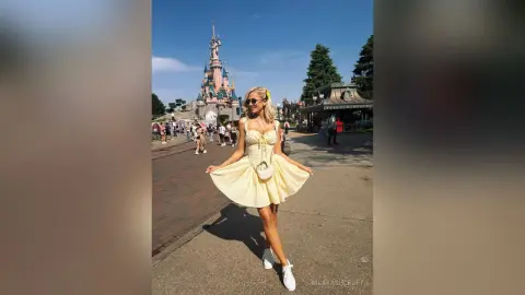 NickyAshcroft poses in a short, light yellow sundress and sunglasses while standing on a paved path with a castle in the background.