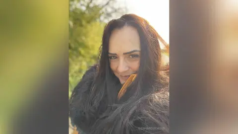 Smiling woman with long dark hair wearing a cozy dark fur coat in a sunny outdoor natural setting.
