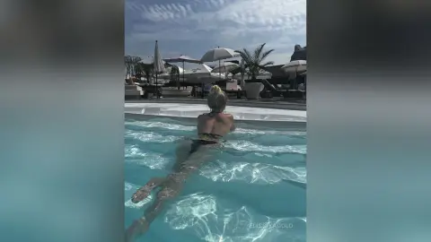 Blonde woman in a black bikini relaxing in a luxury resort swimming pool under a sunny sky.