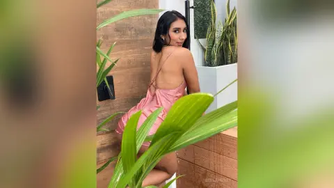 Young woman in a pink satin slip dress posing outdoors near tropical green plants in a sunny setting.