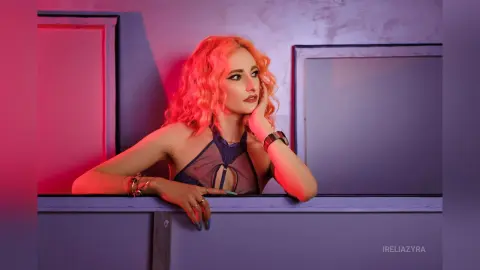 Red-haired woman in a dark mesh top leaning on a window sill in moody pink and purple studio lighting.