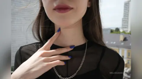Young woman in a black sheer top and silver chain necklace posing outdoors in bright sunlight.