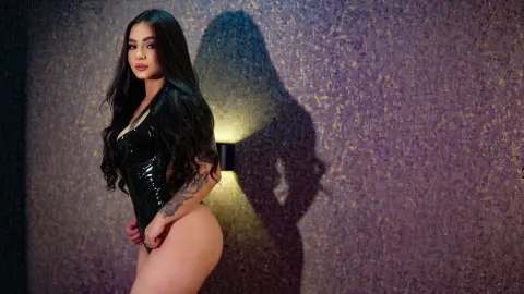Glamorous woman in a black latex bodysuit posing against a textured wall with moody dramatic lighting.