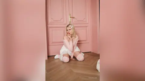 Blonde woman in a dreamy white lace outfit and pink socks sitting on a floor against a soft pink door.