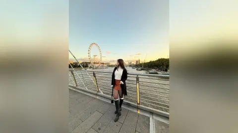 Young woman in a black cardigan and mini skirt posing by the London Eye at sunset.
