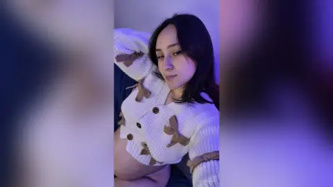 AnnieBuny — Young woman in a cozy white ribbed cardigan with brown bows posing in soft purple lighting.