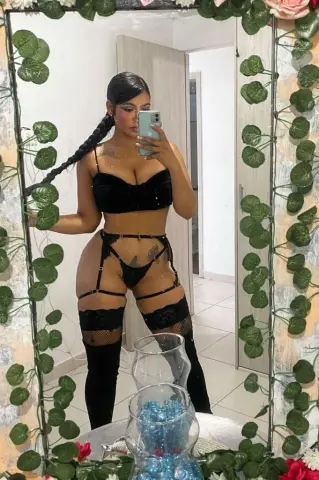 Woman in black lace lingerie and thigh-high stockings taking a mirror selfie in a floral decorated room.