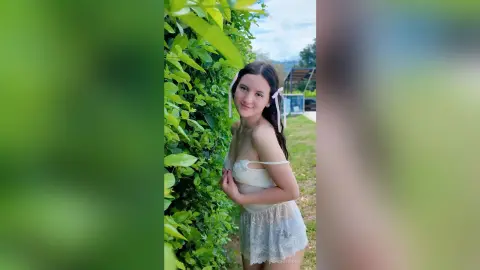Smiling young woman in a white lace sundress standing by a lush green garden hedge on a sunny day.