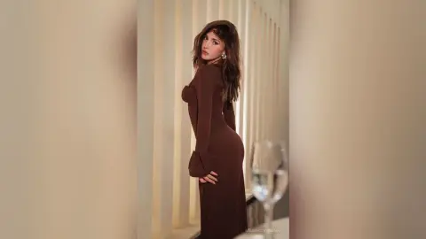 Elegant woman in a long brown dress posing in a bright, minimalist indoor setting with a sophisticated mood.