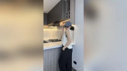 Stylish woman in a white tank top and black trousers posing in a modern minimalist kitchen.