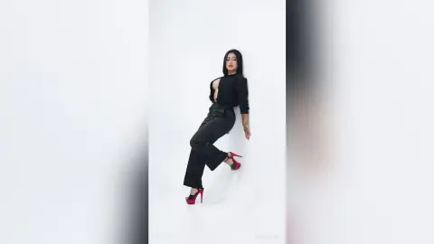 KimRoberth poses in black trousers and a black long-sleeved crop top with red high heels against a white background.