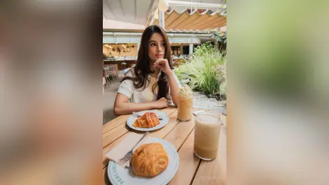 TefyRusso sits at an outdoor cafe table with long brown hair, wearing a white short-sleeved top next to plates of pastries and iced coffee.