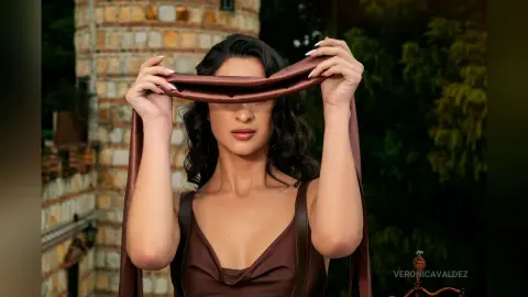 VeronicaValdez holds a brown silk strap over her eyes, wearing a brown tank top with her dark wavy hair down.