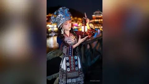 Young woman in traditional silver ethnic costume holding a lantern at night near a glowing village.