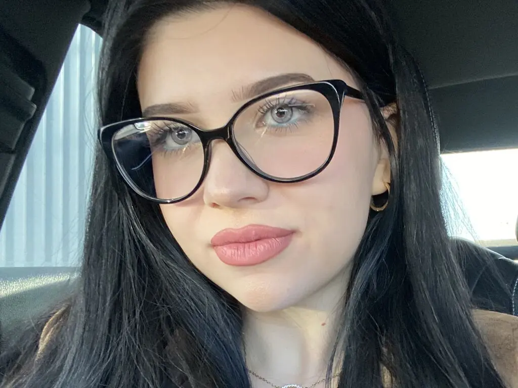 Young woman with black hair and black glasses smiling softly in a car setting.