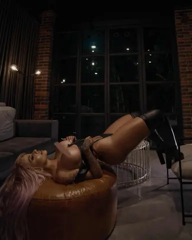 LucyHills39 lies on a brown leather ottoman, wearing black lingerie and thigh-high boots, showing her long pink hair and toned body in a dark room.