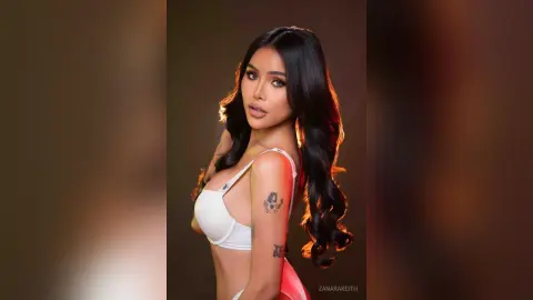 Glamorous woman in a white bikini top posing with long wavy hair against a warm studio background.