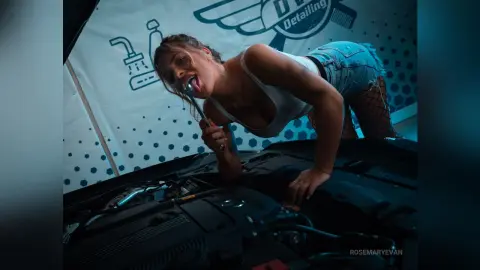 Playful woman in a white tank top and denim shorts leaning over a car engine in a blue detailing garage.