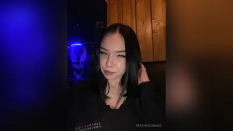 Young woman with long dark hair wearing a black graphic t-shirt in a moody, dimly lit room with blue neon lighting.