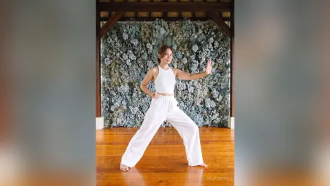 MilaSwan — MilaSwan poses in a white crop top and wide-leg pants against a blue floral wall background.