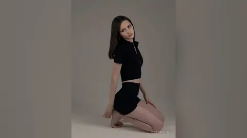 Young woman in a black short-sleeve crop top and mini skirt posing against a minimalist grey studio background.
