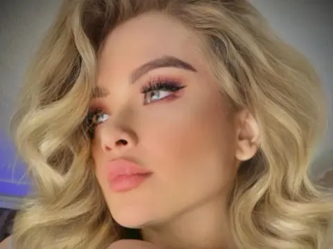 Close-up portrait of a blonde woman with glamorous makeup and wavy hair in soft indoor lighting.