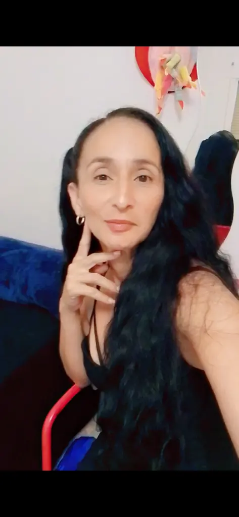 Woman with long curly hair in a black strapless corset sitting near a green houseplant in warm indoor lighting.