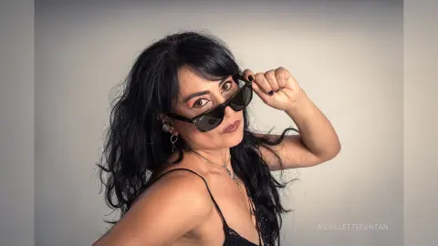 Stylish woman with long dark hair wearing sunglasses and a black strap outfit against a neutral studio background.