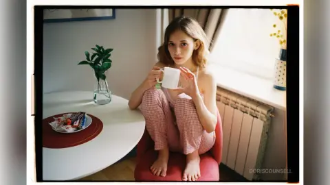 Young woman in pink checkered pajamas sitting at a table with a coffee mug in a bright, cozy morning setting.