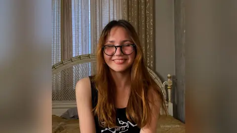 Smiling young woman with long brown hair and glasses wearing a black tank top in a cozy bedroom setting.