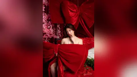 Elegant woman in a red velvet outfit sitting amidst pink cherry blossoms in a romantic red setting.