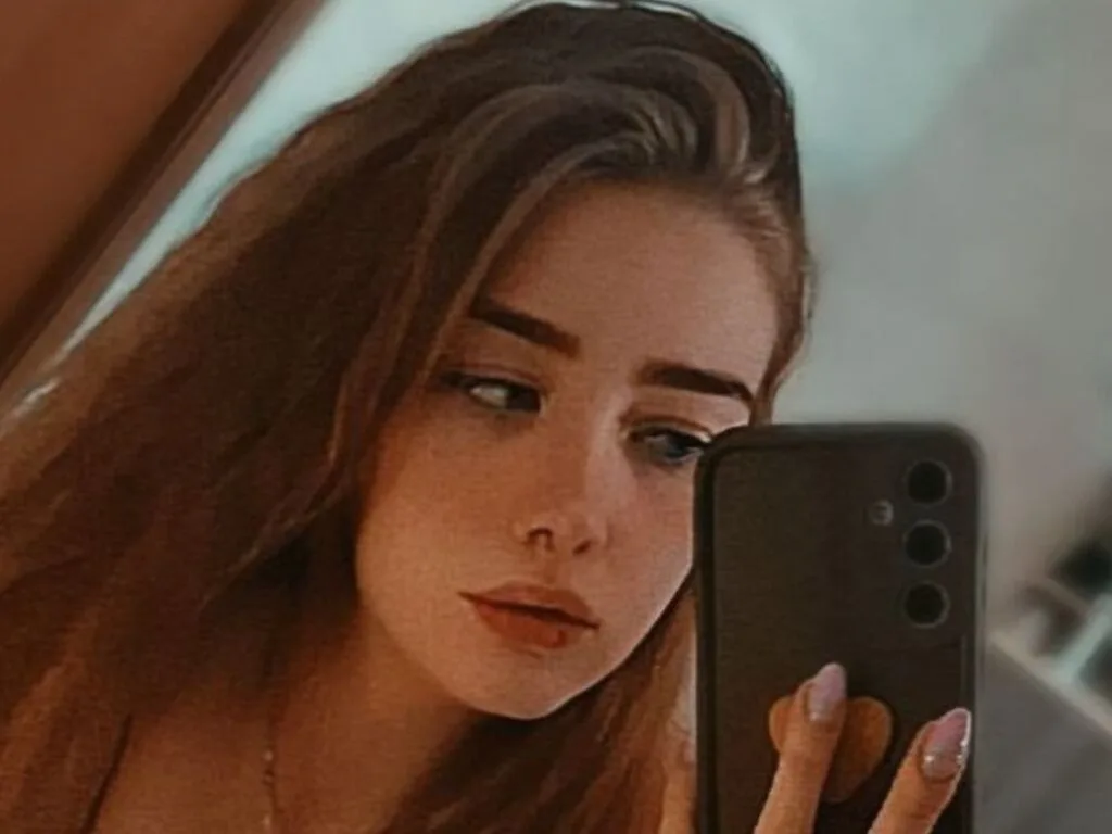 TeresiaCaspi takes a mirror selfie with her phone, looking into the camera with long brown hair.