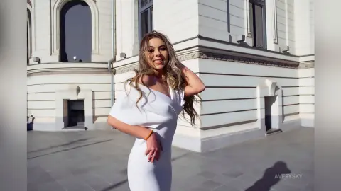 Smiling woman in a white elegant dress posing in front of a classic white building on a sunny day.