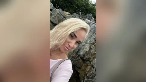 Blonde woman in a white top smiling in front of a scenic rocky waterfall outdoors.